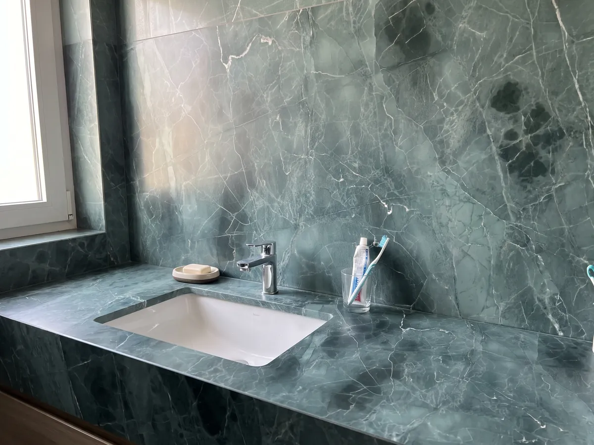 Cubierta de lavabo y pared de baño en mármol verde Green Algae con lavamanos blanco y grifería cromada: verde azulado con nubes más oscuras y una red de vetas blancas finas