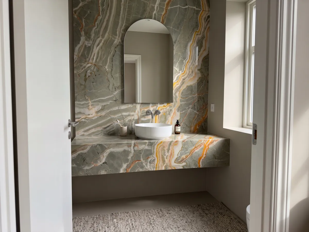 Cubierta de lavabo y pared de baño en ónix verde Rainbow con lavamanos blanco y grifería cromada: verde grisáceo cruzado por vetas naranja y amarillas ramificadas