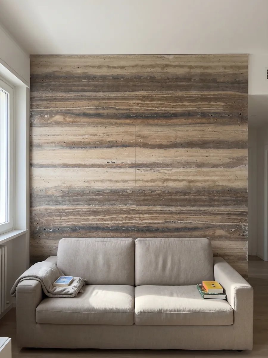 Pared de sala revestida con placas de travertino marrón Oscuro Silver abiertas en libro detrás de un sofá: franjas horizontales marrones y beige muy marcadas a lo largo de la placa