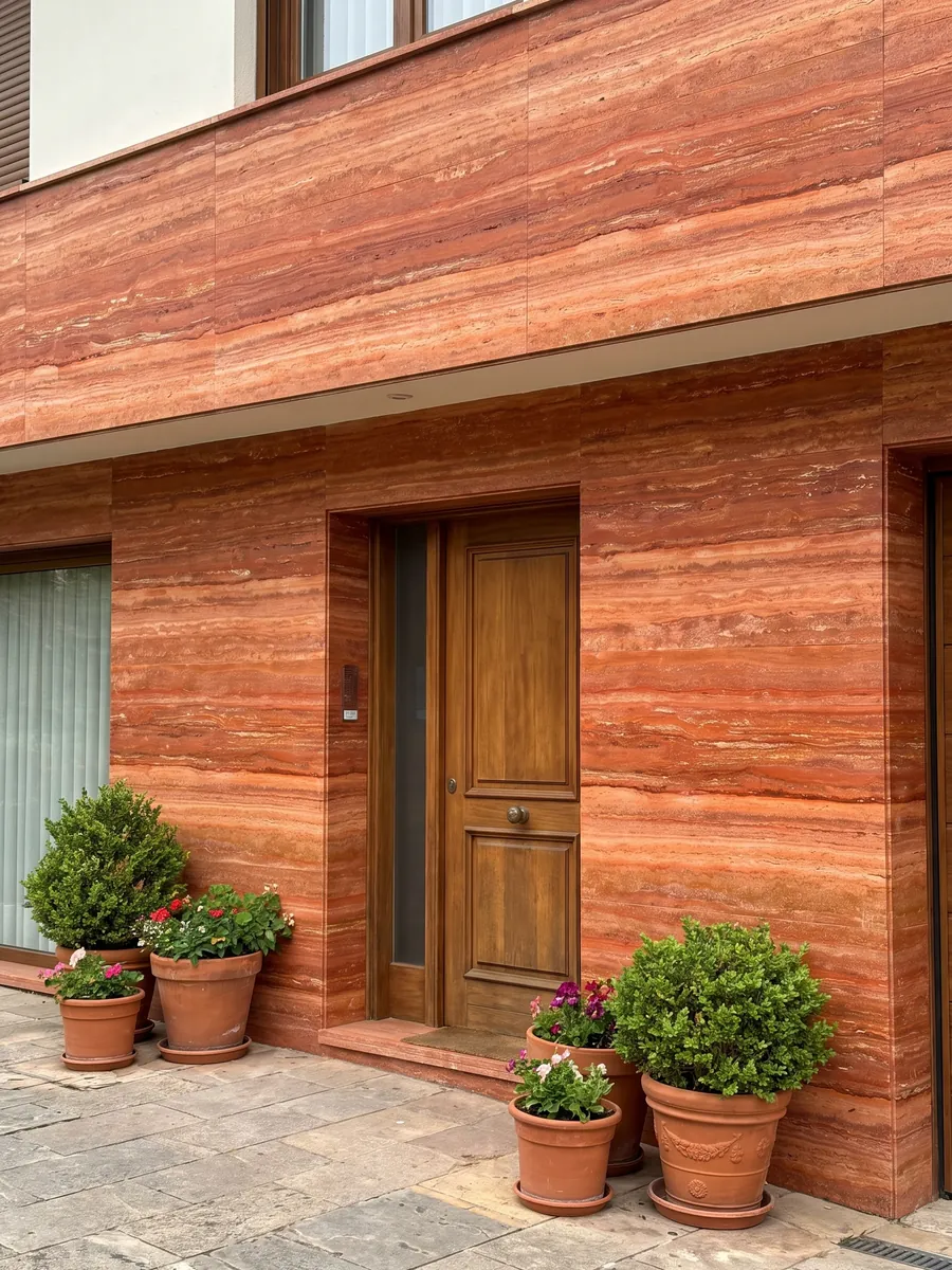 Fachada de casa revestida con Travertino Rojo junto a una puerta de madera: rojo anaranjado intenso en franjas horizontales con líneas crema finas