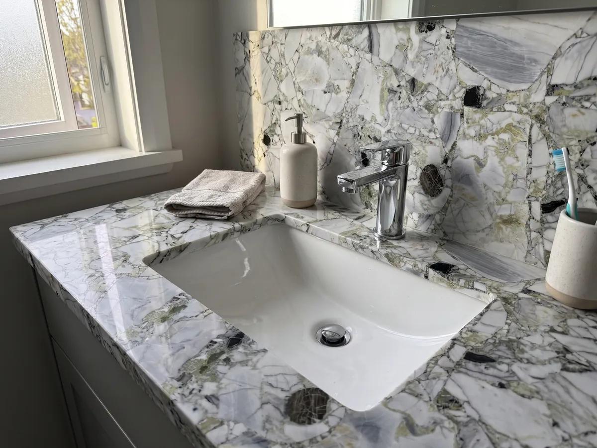 Cubierta de lavabo y pared de baño en White Beauty con lavamanos blanco y grifería cromada: fragmentos blancos y grises unidos por zonas verde claro, con piezas negras sueltas