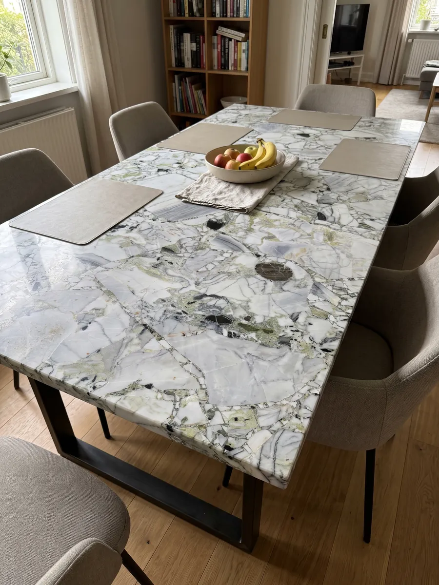 Mesa de comedor con cubierta de White Beauty sobre base de acero: fragmentos blancos y grises unidos por zonas verde claro, con piezas negras sueltas
