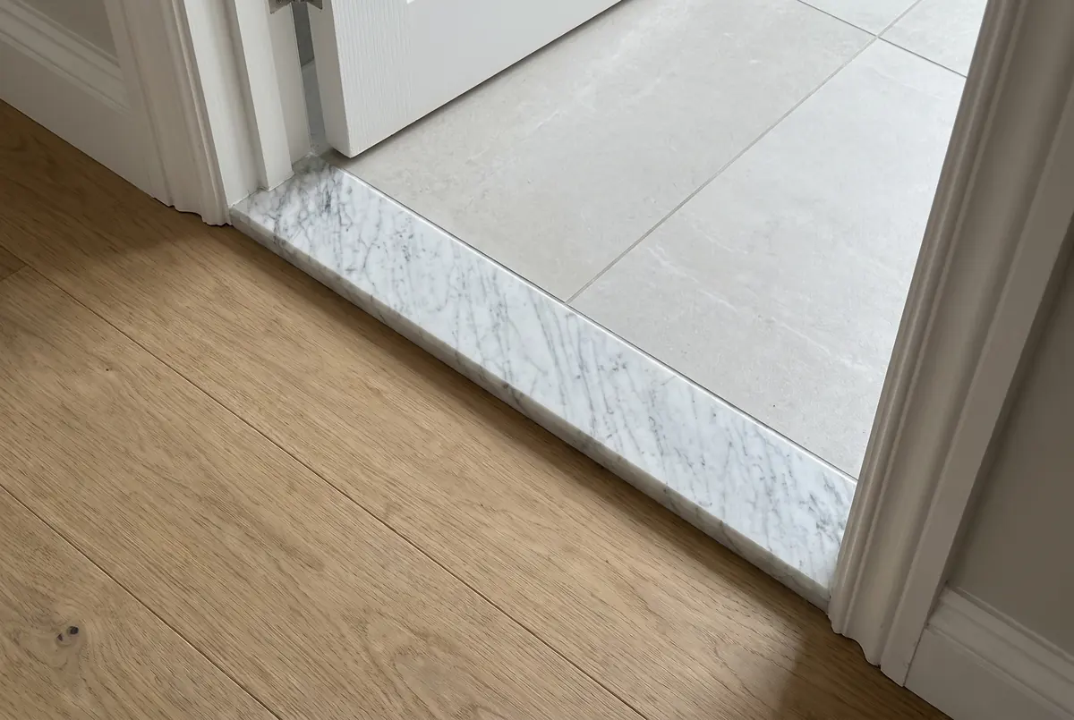 Umbral de mármol blanco con vetas grises en el paso de una puerta, entre un piso de madera y uno de baldosa clara